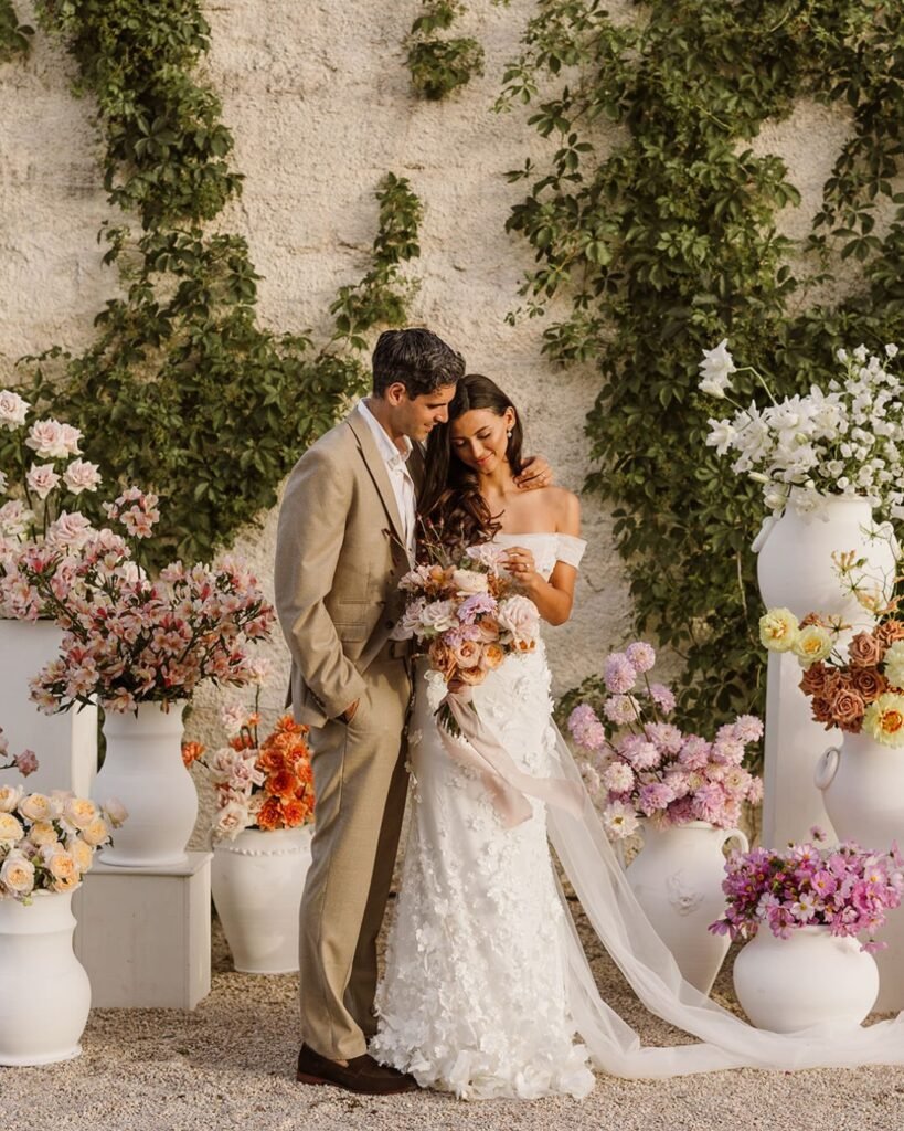 Bride and groom standing among lush floral arrangements at Masseria Fontana di Vite, surrounded by soft pastels and greenery.
