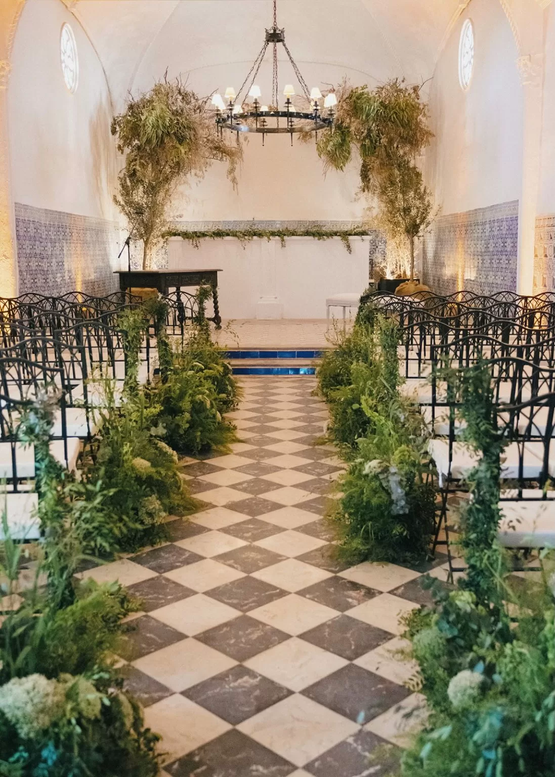 Indoor wedding ceremony aisle with lush greenery and checkered floor at Masia Aldamar