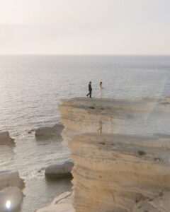 A newlywed couple stands on a rocky cliff overlooking the Mediterranean Sea, captured by Karina Leonenko.