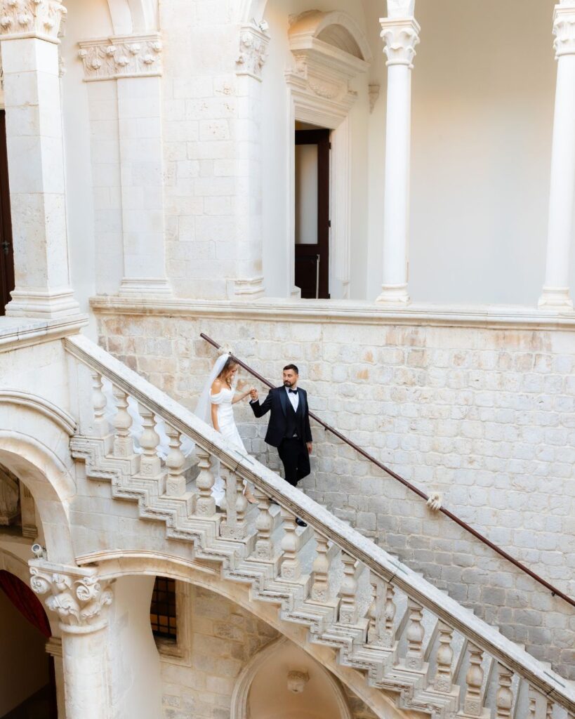 The bride and groom walk down a grand staircase together, the elegance of the moment captured by Kristijan Rezo.