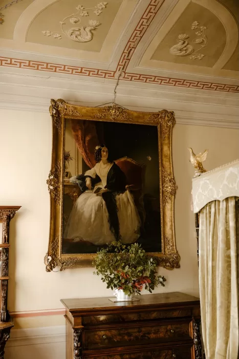 Grand oil portrait of a lady in white, framed in gold above a dark wood chest in Borgo Stomennano’s historic bedroom.