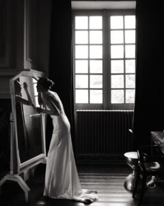 Bride applying makeup at an antique mirror near a tall window at Château Rigaud, captured in quiet focus.