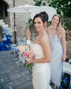 The bride smiles under a white parasol at Castello Montignano, holding a pastel bouquet of peonies, delphiniums, and roses, with her bridesmaid behind her.