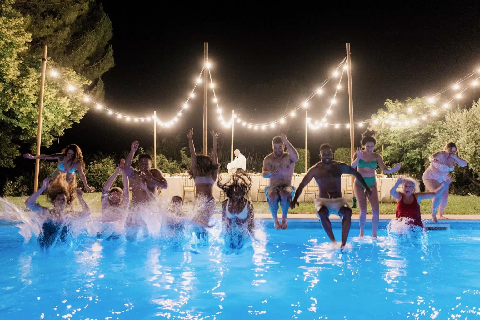 Wedding guests jump into the glowing blue pool under string lights during a lively night celebration at Castello di Montignano.