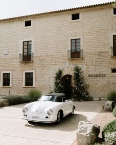 Classic white wedding car parked in front of the historic stone facade and floral-decorated entrance of Finca Morneta.
