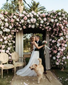 The couple kisses under a lush flower arch as their dog walks past mid-celebration — joyful candid moment by Lima de Miguel.