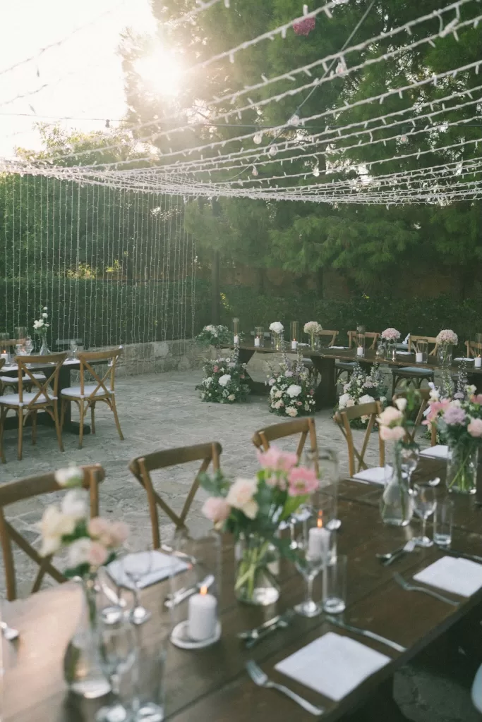 Outdoor reception setup at Liopetro with hanging fairy lights and floral arrangements glowing in the golden hour sun.
