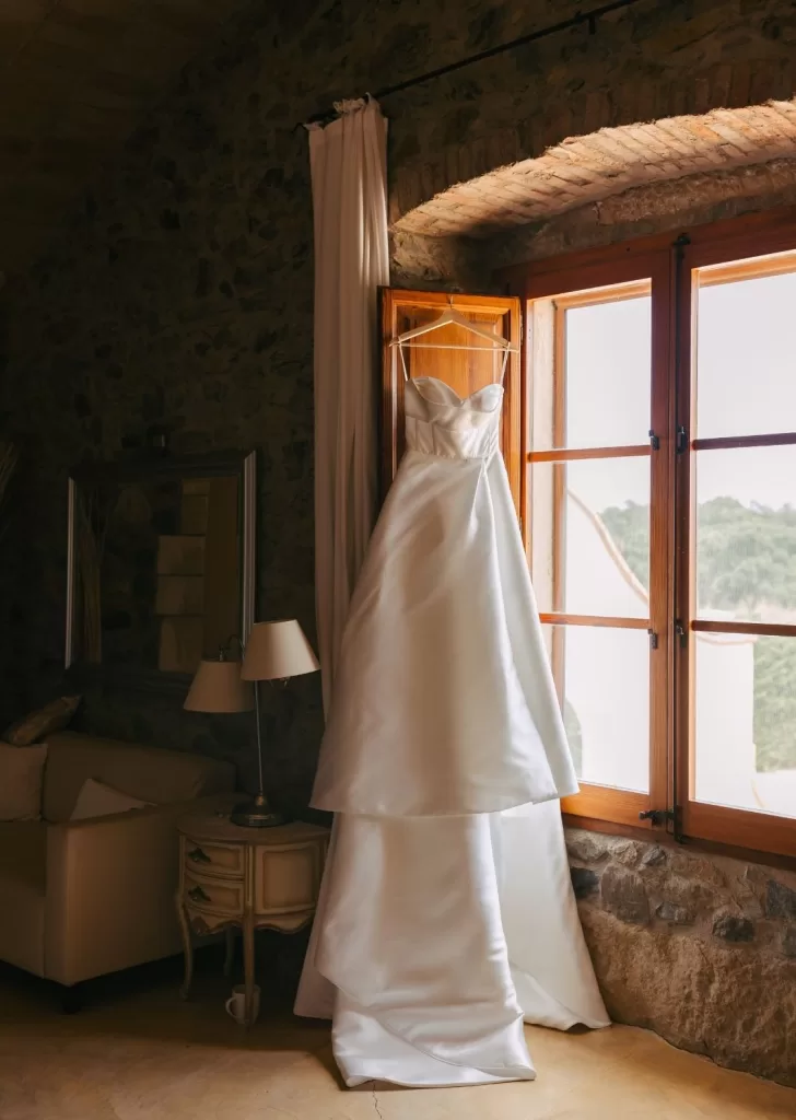 Strapless satin wedding gown hanging in a sunlit stone room window at Mas Torroella