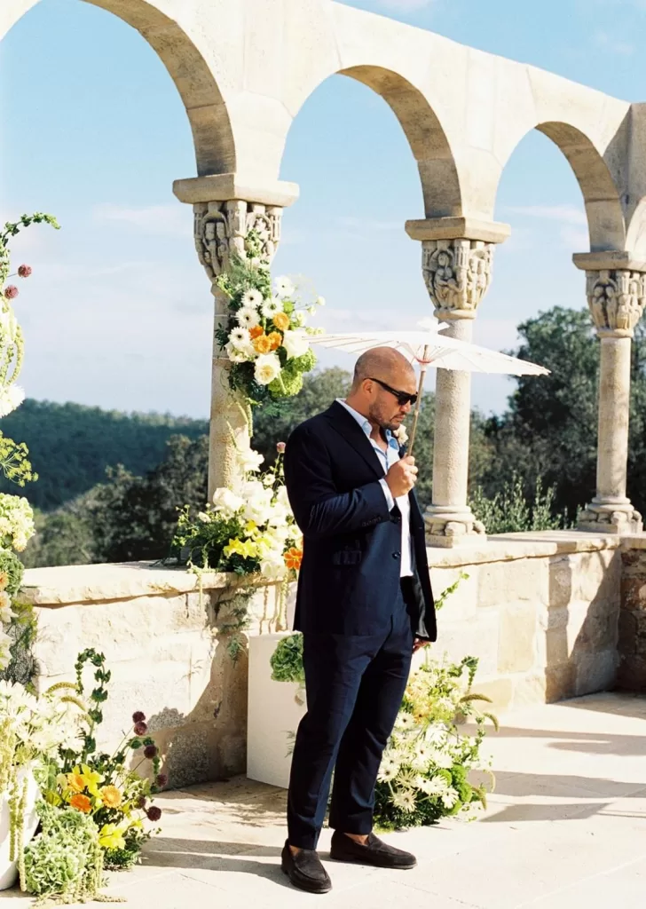 Groom holding a parasol, standing beneath the historic archway at Mas Torroella, surrounded by floral arrangements.