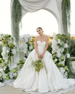 Bride in voluminous gown under romantic floral arch with ocean in background, photographed by Puy Cermeño.