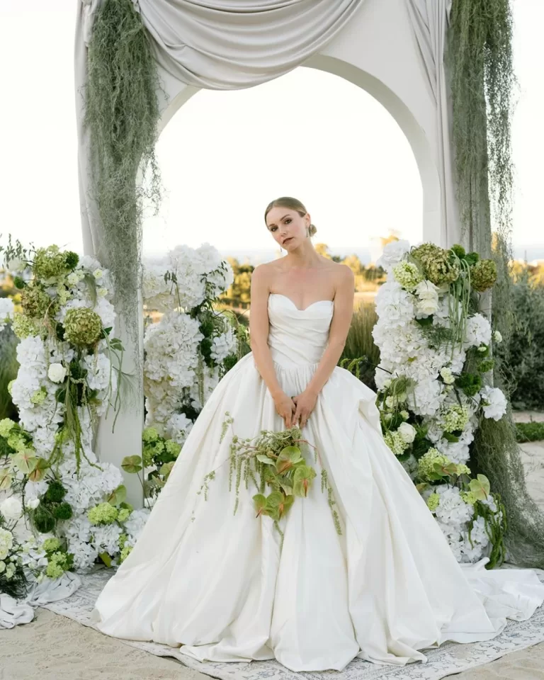 Bride in voluminous gown under romantic floral arch with ocean in background, photographed by Puy Cermeño.