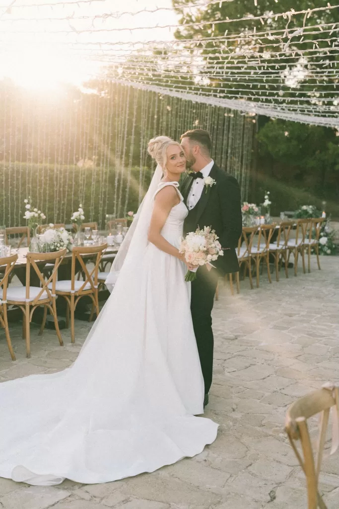 Groom kisses his bride’s temple as she smiles, wrapped in golden-hour light at Liopetro’s outdoor reception.