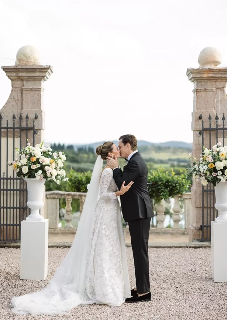 Bride and groom kiss at the altar framed by floral urns and stone pillars at Villa Cariolla, with rolling hills in the background.