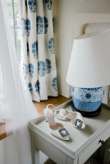 Elegant bridal shoes placed on a bedside table near floral curtains and a blue ceramic lamp.