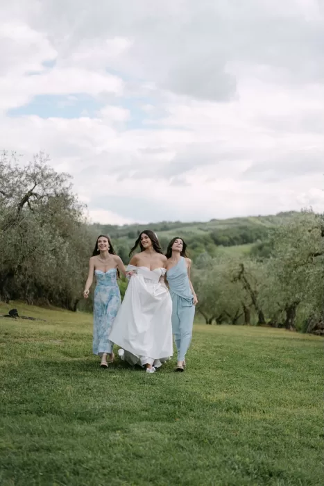 The bride and her bridesmaids walking through an olive grove at Tenuta Corbinaia, sharing a lighthearted moment.
