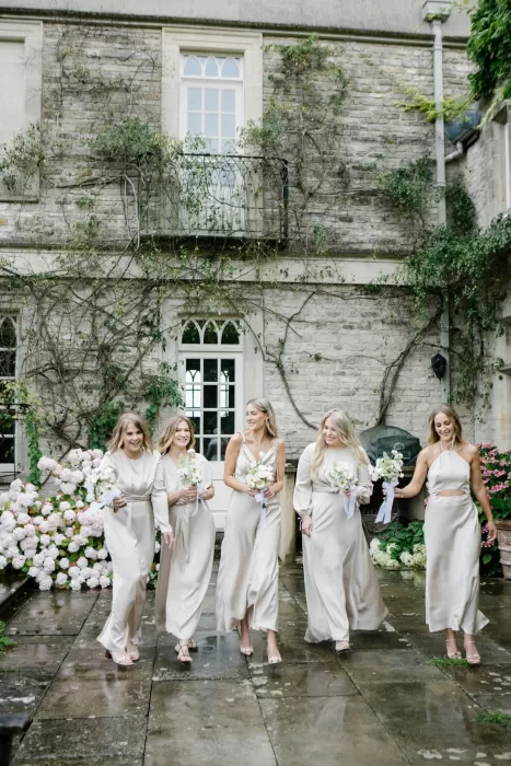 Bridesmaids in ivory dresses smiling and walking through the courtyard at Euridge Manor holding white bouquets.