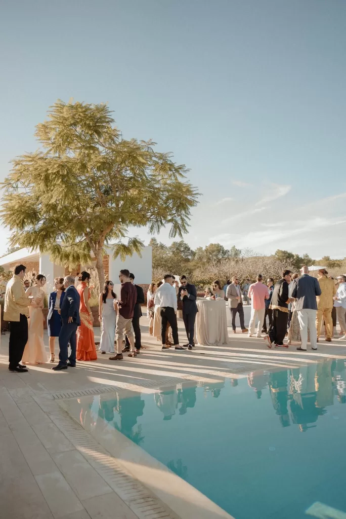 Wedding guests mingling by the pool during cocktail hour at Ca Na Xica, surrounded by warm light and trees.
