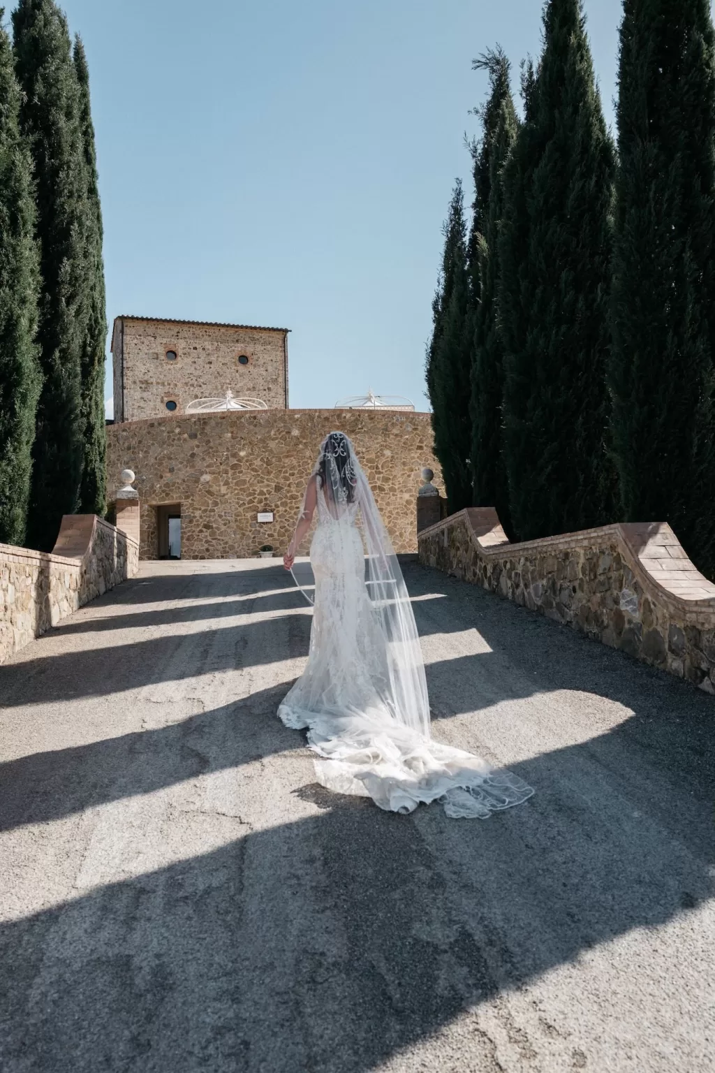 Bride in lace gown walking up the stone pathway lined with cypress trees at Castello di Velona.
