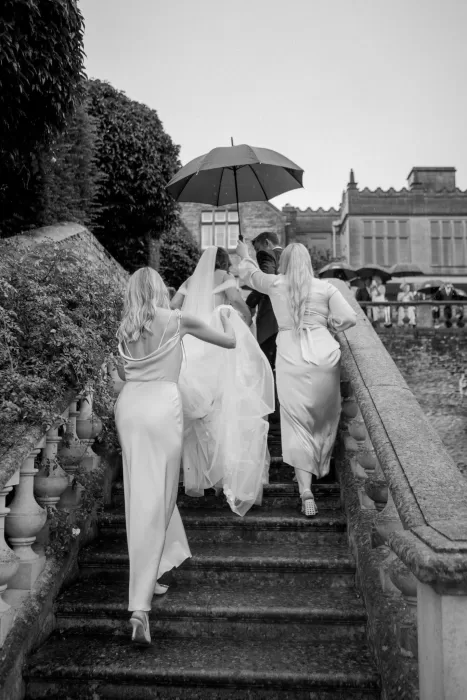 The bride walks up the stone steps of Euridge Manor under an umbrella, flanked by bridesmaids in satin gowns.