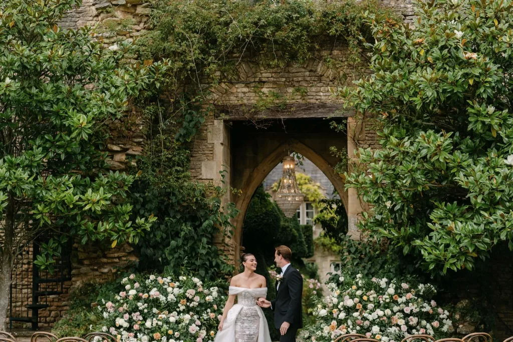 Bride and groom holding hands in front of Euridge Manor’s romantic stone archway and abundant ceremony florals.