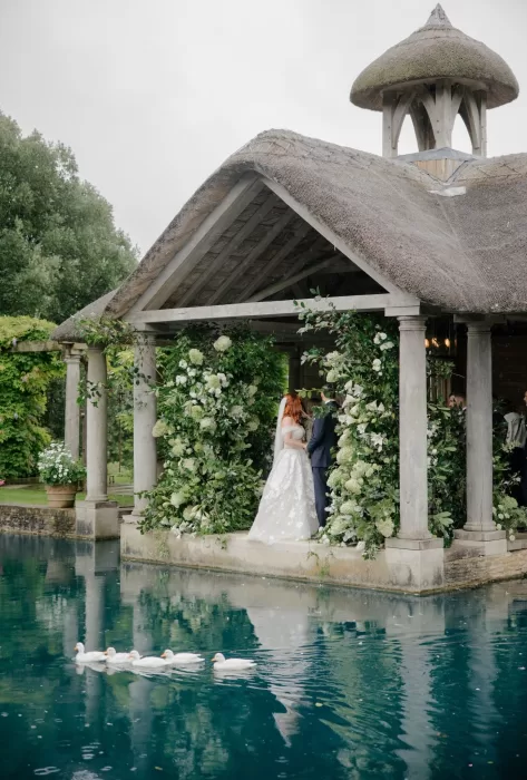 The couple exchanges vows beneath the pavilion at Euridge Manor, while a line of ducks glides across the water.