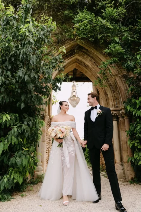 Couple walking hand-in-hand beneath the ivy-covered gothic archway at Euridge Manor with a crystal chandelier above.