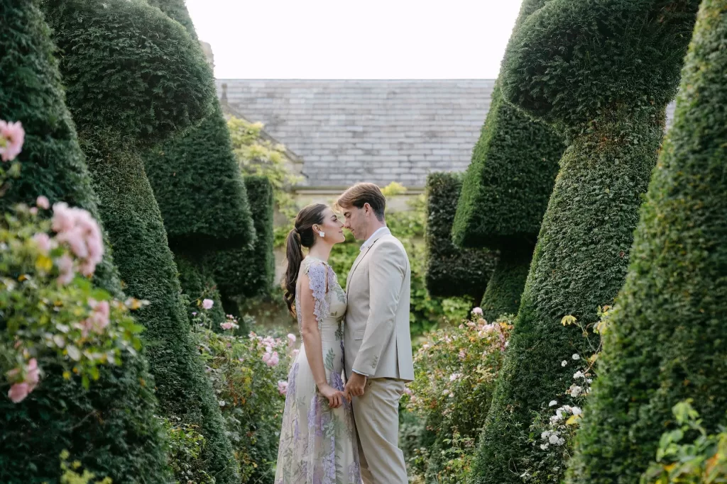 Couple standing between sculpted topiary hedges and blooming roses at Euridge Manor, surrounded by garden serenity.