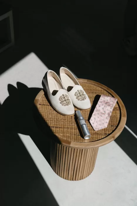 Groom’s embroidered loafers, tie, and perfume artfully styled on a woven side table at Masia Casa del Mar.
