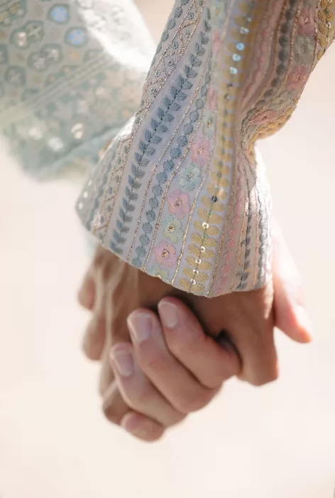 Close-up of pastel floral embroidery on a wedding sherwani, with two grooms gently holding hands at Masia Casa del Mar.