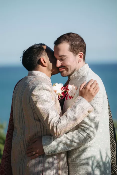 Emotional embrace between two grooms in pastel attire against a coastal backdrop at Masia Casa del Mar.