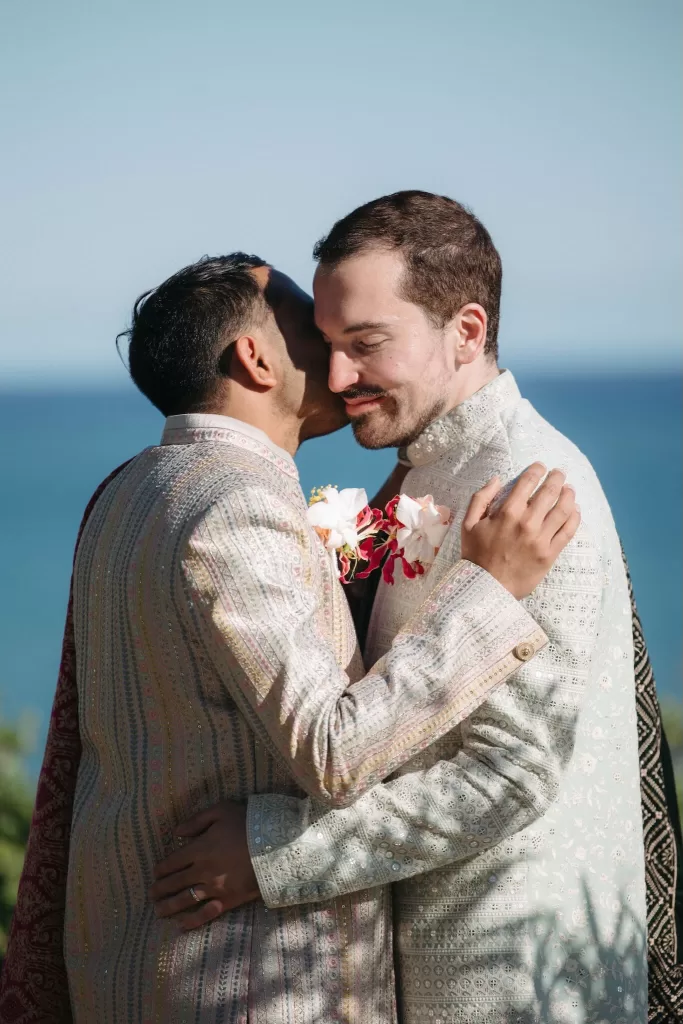 Emotional embrace between two grooms in pastel attire against a coastal backdrop at Masia Casa del Mar.