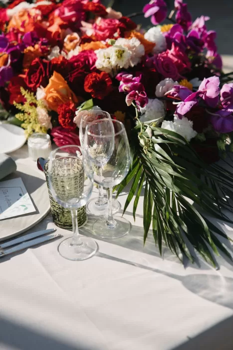 Lush, vibrant floral arrangements of roses and orchids adorn a reception table at Masia Casa del Mar, with sunlight glinting off glassware.