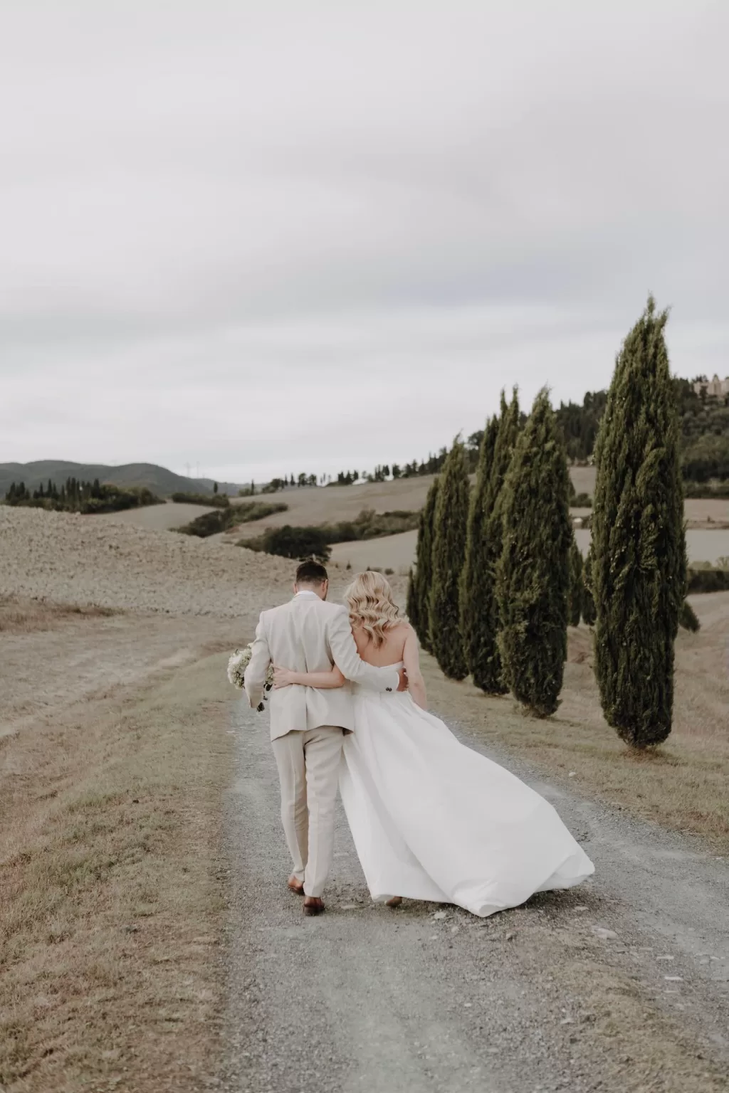 Bride and groom embrace while walking down a gravel path lined with cypress trees at Podere Calvaiola.