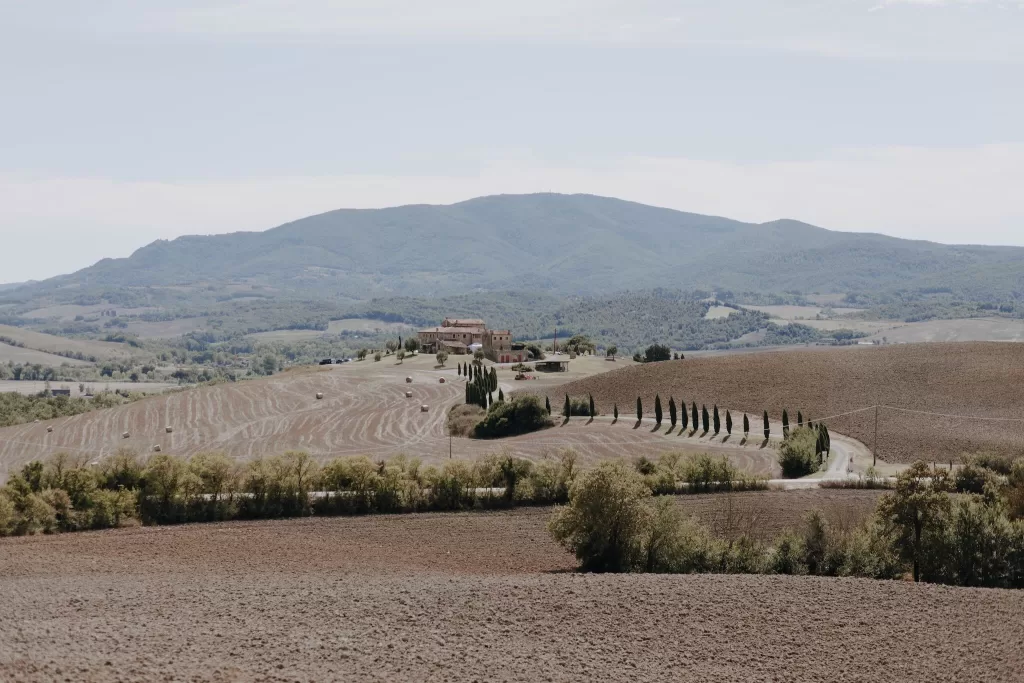 Panoramic view of Podere Calvaiola with winding cypress-lined road and Tuscan hills in the distance.