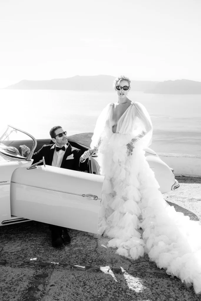 Bride in dramatic tulle gown stands beside groom in classic tuxedo and vintage car with the Aegean Sea behind them at Rocabella Santorini.