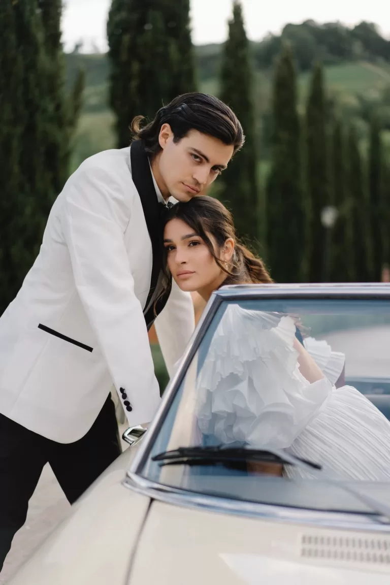 Intimate moment between bride and groom leaning against a vintage car surrounded by Tuscan cypress trees at Tenuta Corbinaia.