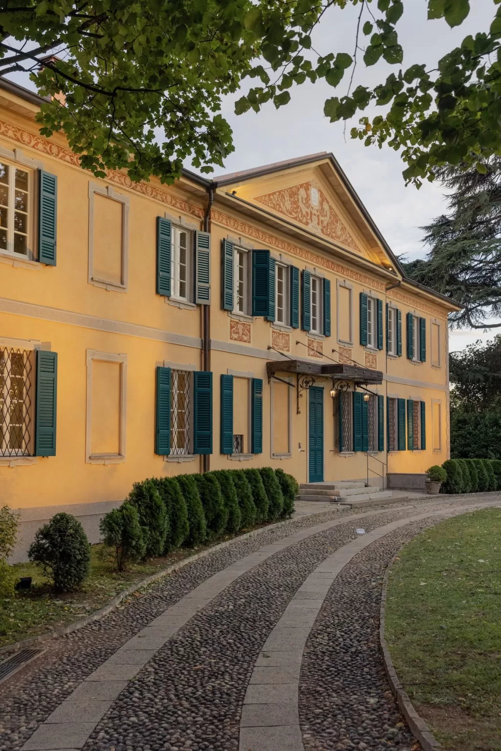 Curved cobblestone path leads to the yellow façade and teal shutters of Villa Buttafava, bathed in evening light.