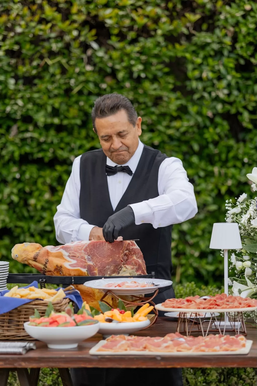 A chef in formal attire slices cured ham at an elegant outdoor catering station in Villa Buttafava’s garden.