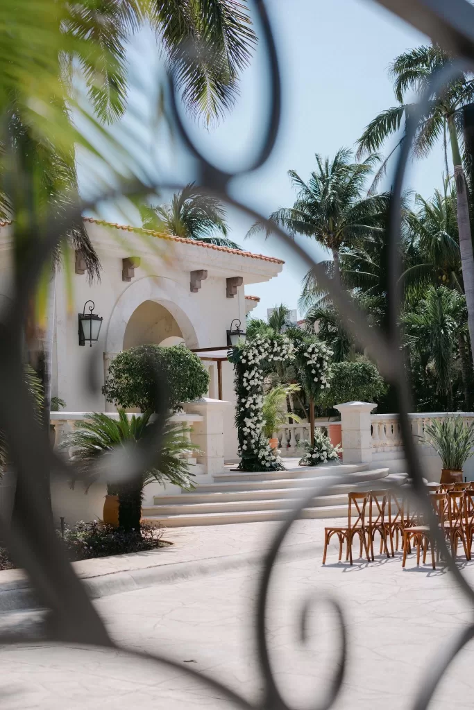 Ceremony space at Villa La Joya seen through wrought iron detail, framed by palms and floral arch.