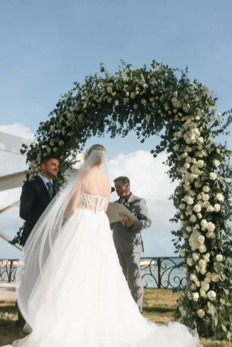 Bride and groom exchange vows beneath a floral arch with ocean backdrop at Villa La Joya.