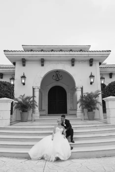 Couple sits on grand front steps beneath the arched entrance of Villa La Joya