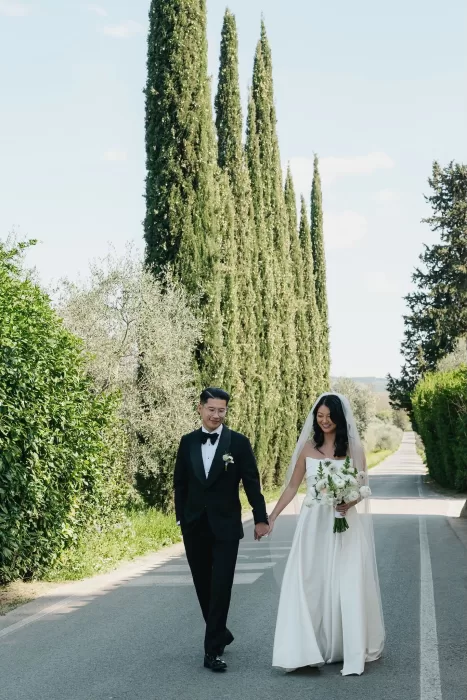 Bride and groom walking hand in hand along a tree-lined road near Villa Medicea di Lilliano, surrounded by cypress trees.