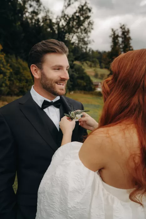 Bride pins a delicate boutonniere on the groom’s lapel as they smile under overcast skies at Hotel Villa Honegg.