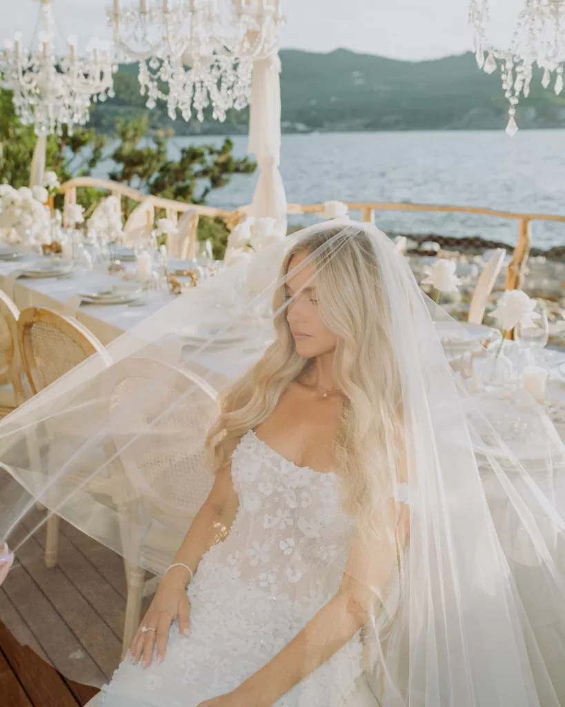 The bride sits at the elegantly styled dinner table with chandeliers above, her veil softly draped over her face.