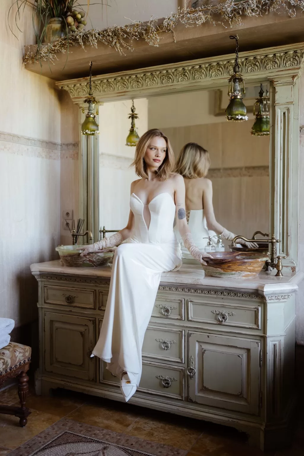 Bride in strapless gown sitting on vintage dresser with mirror in Chateau Challain suite.