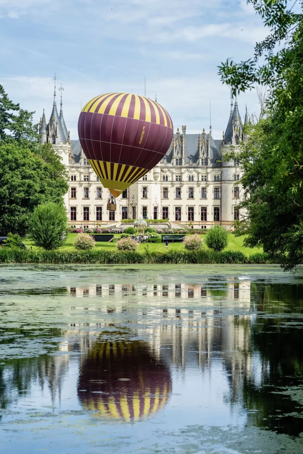 Striped hot air balloon hovering in front of Château Challain with its reflection mirrored in the château’s pond.