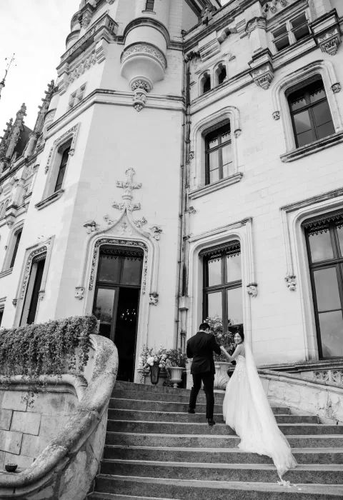Groom helping bride up the staircase of Château Challain, framed by gothic architecture.