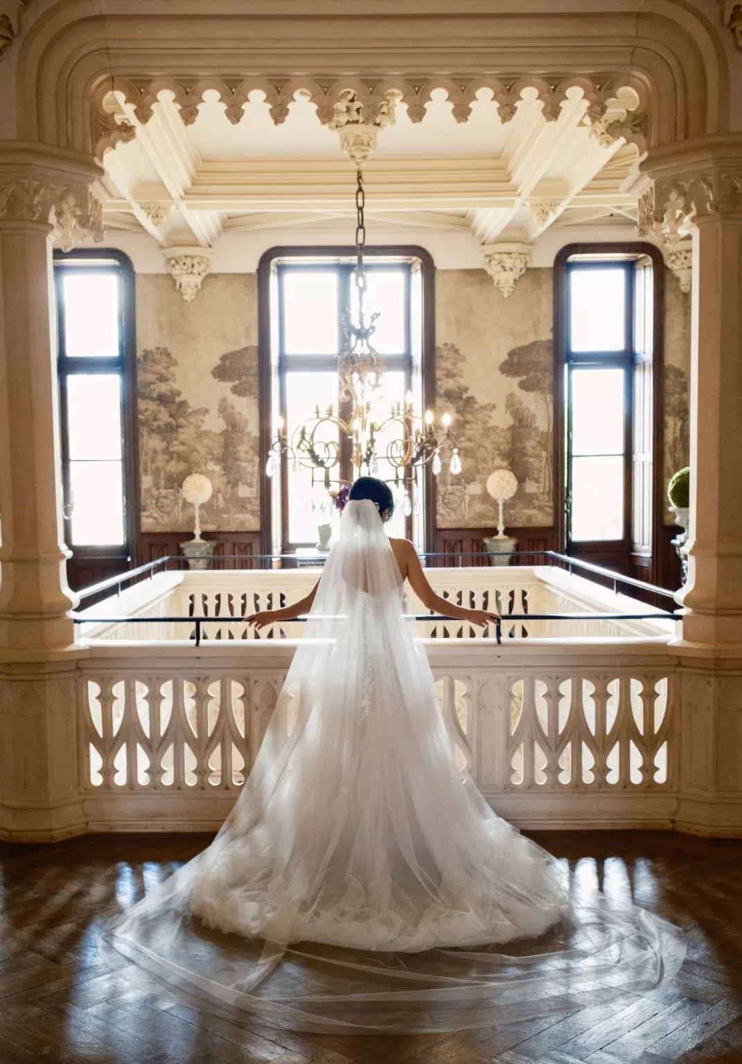 Back view of the bride on the grand staircase at Château Challain, framed by gothic arches and chandeliers.