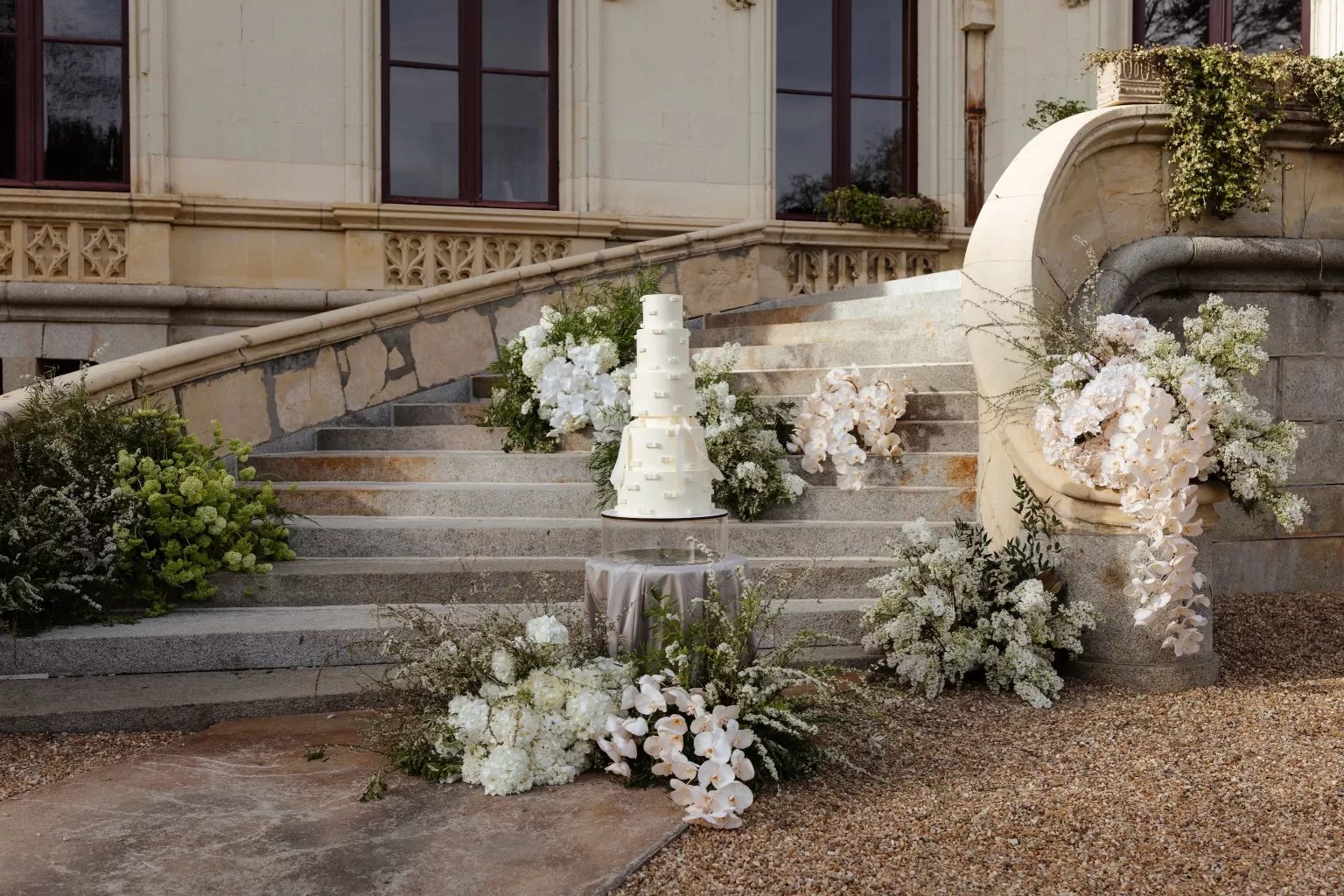 Elegant tiered wedding cake surrounded by cascading white florals on the stone steps of Chateau Challain.