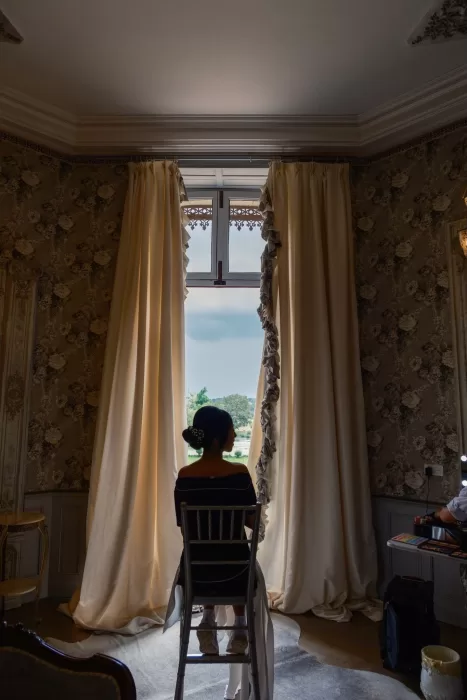 Bride sits in front of a tall window at Château Challain, framed by elegant curtains and soft daylight.